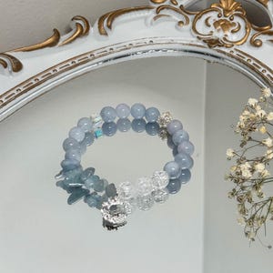 May include: A beaded bracelet featuring light blue and clear beads, accented with silver-colored embellishments. The bracelet is displayed on a reflective surface, showcasing its design and details. The bracelet is a piece of jewelry.