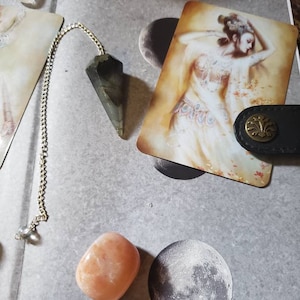 Op de afbeelding: Een groene kristallen slinger aan een zilveren ketting, een licht oranje steen en een kaart met een vrouw in een witte jurk en een maan op de achtergrond.
