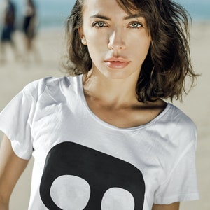 Op de afbeelding: Witte crop top met een grote zwarte schedelafbeelding. De top heeft een ronde hals en korte mouwen. Het model staat op een strand met de oceaan op de achtergrond.