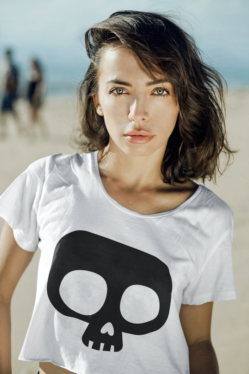 Op de afbeelding: Witte crop top met een grote zwarte schedelafbeelding. De top heeft een ronde hals en korte mouwen. Het model staat op een strand met de oceaan op de achtergrond.
