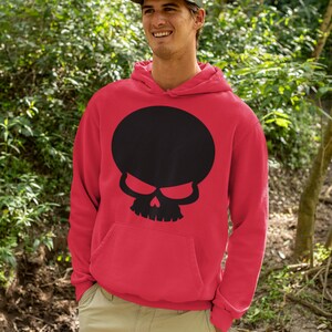 Op de afbeelding: Rode hoodie met een grote zwarte schedelafbeelding. Het schedelontwerp heeft een ronde hoofdvorm en dreigende ogen. De hoodie heeft een voorzak en wordt gedragen met een groene pet en een kaki broek.