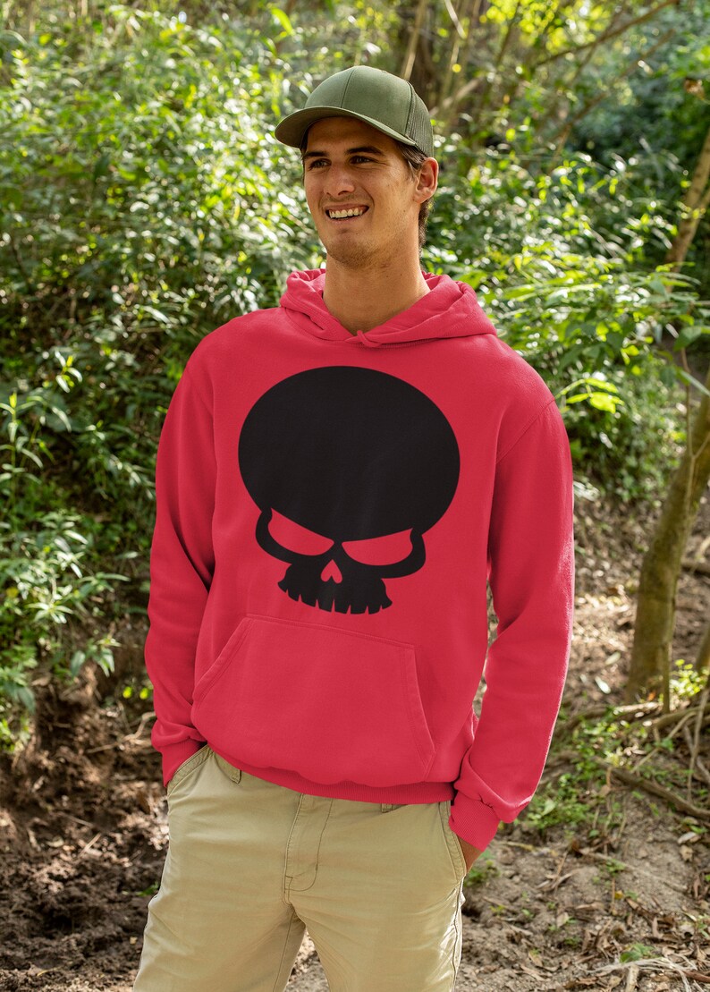 Op de afbeelding: Rode hoodie met een grote zwarte schedelafbeelding. Het schedelontwerp heeft een ronde hoofdvorm en dreigende ogen. De hoodie heeft een voorzak en wordt gedragen met een groene pet en een kaki broek.