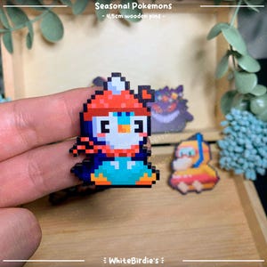 May include: A set of four 4.5cm wooden pins featuring pixel art designs of Pokemon characters. The pins are of a penguin wearing a red hat, a black and white creature with a red eye, and a yellow character with a blue helmet. The text "Seasonal Pokemons" is at the top of the image.