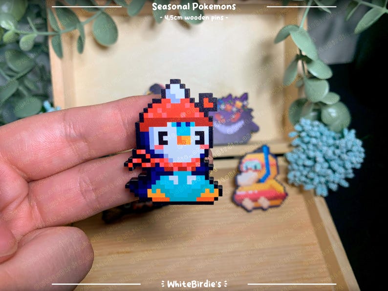 May include: A set of four 4.5cm wooden pins featuring pixel art designs of Pokemon characters. The pins are of a penguin wearing a red hat, a black and white creature with a red eye, and a yellow character with a blue helmet. The text "Seasonal Pokemons" is at the top of the image.