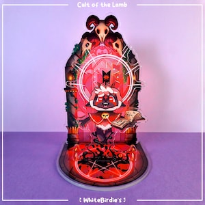 May include: A clear acrylic standee featuring a cartoon lamb character from "Cult of the Lamb." The lamb wears a red cloak and holds a book. The standee has a red and black color scheme with the text "Cult of the Lamb" at the top.