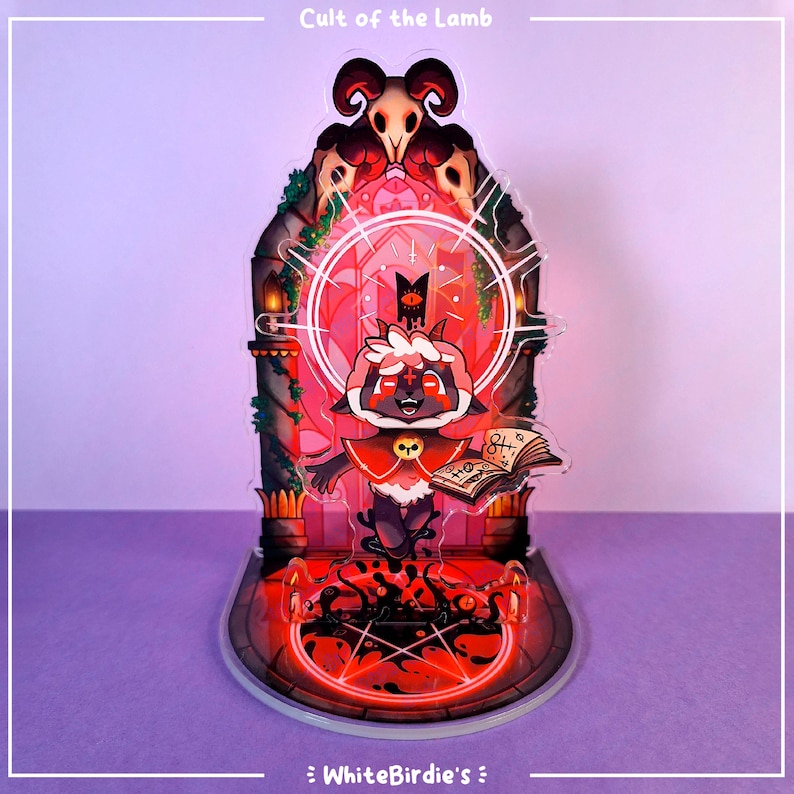 May include: A clear acrylic standee featuring a cartoon lamb character from "Cult of the Lamb." The lamb wears a red cloak and holds a book. The standee has a red and black color scheme with the text "Cult of the Lamb" at the top.
