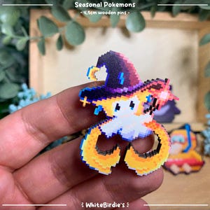 May include: A wooden pin featuring a pixelated Pokemon character wearing a purple and black wizard hat. The character has yellow and orange fur and is holding a wand. The pin is 1.5 inches in diameter.
