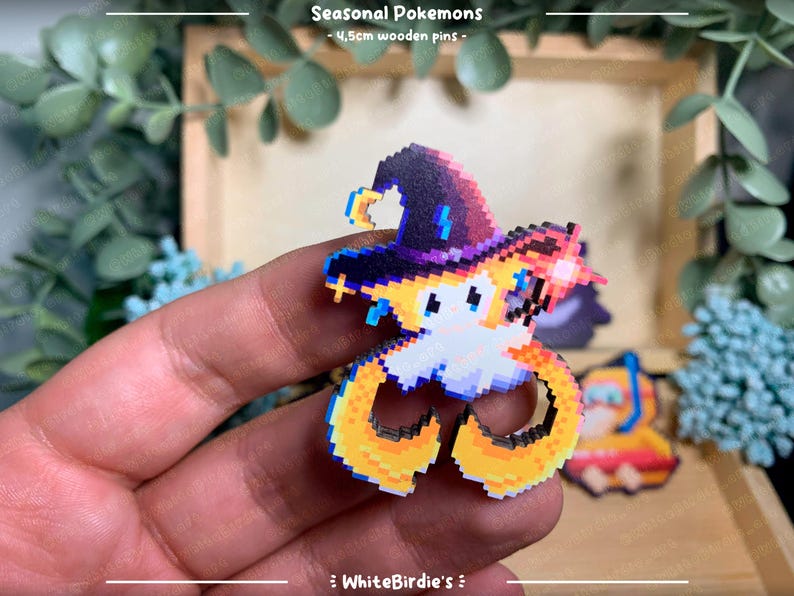May include: A wooden pin featuring a pixelated Pokemon character wearing a purple and black wizard hat. The character has yellow and orange fur and is holding a wand. The pin is 1.5 inches in diameter.