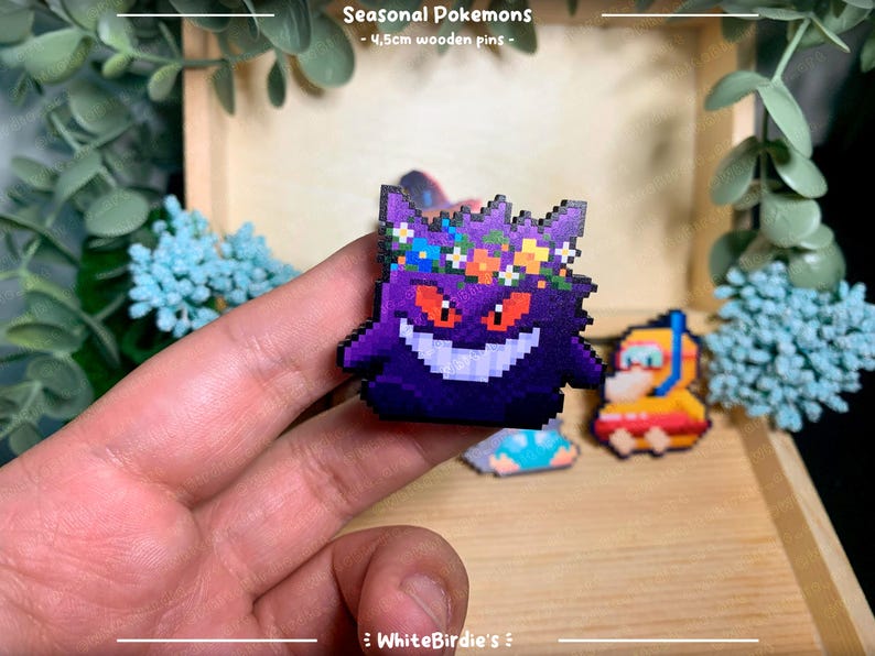 May include: A purple and black Gengar Pokemon pin with a flower crown. The pin is made of wood and is approximately 4.5cm in size. The pin is part of a collection of seasonal Pokemon pins.