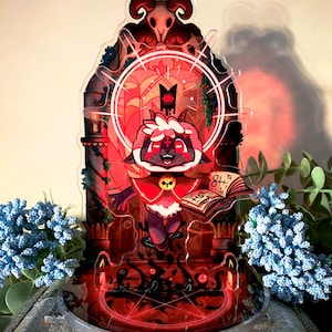 May include: A decorative acrylic pin featuring a cartoon lamb character with a red and white outfit, holding a book. The pin is set against a red and black background with the text "Cult of the Lamb". The pin is 4.5cm.