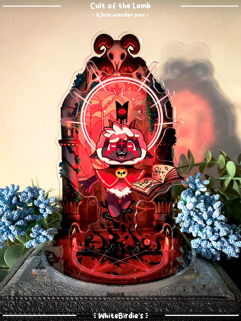 May include: A decorative acrylic pin featuring a cartoon lamb character with a red and white outfit, holding a book. The pin is set against a red and black background with the text "Cult of the Lamb". The pin is 4.5cm.