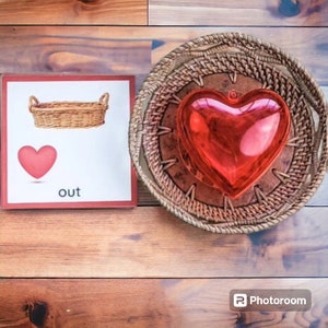 May include: A red heart-shaped object is in a woven basket. A red heart is on a white card with the word "out" printed below it. The card is surrounded by a red border.