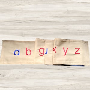 May include: Beige fabric squares with the letters a, b, g, x, y, and z embroidered in red and blue.
