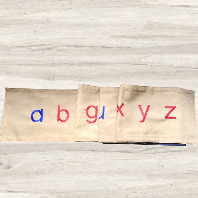 May include: Beige fabric squares with the letters a, b, g, x, y, and z embroidered in red and blue.