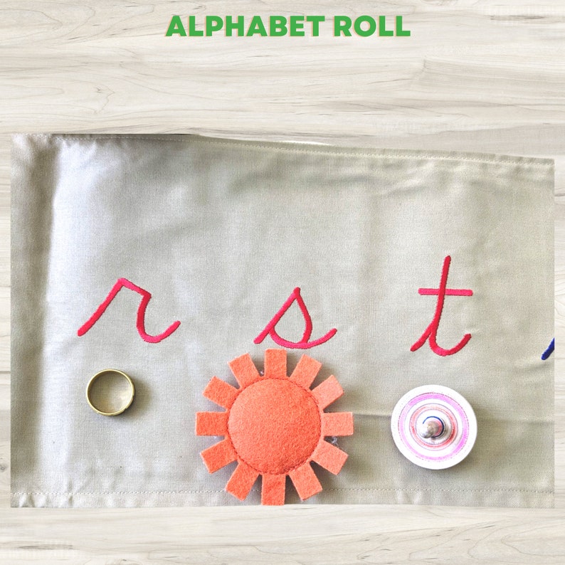 May include: A beige fabric play mat with the letters "r", "s", and "t" embroidered in pink. There is a gold ring, an orange felt sun, and a white and pink spinning top on the mat.