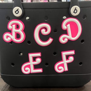 Könnte beinhalten: Schwarze Tragetasche mit weißen und pinkfarbenen Buchstaben, die B, C, D, E und F buchstabieren. Die Buchstaben haben einen weißen Umriss und ein leuchtend pinkfarbenes Innere. Die Tasche hat zwei Griffe und kleine Löcher.