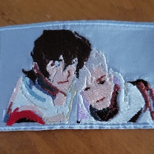 May include: Embroidered patch featuring two anime characters, one with brown hair and the other with white hair, against a light blue background.