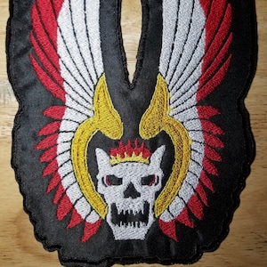 May include: Black embroidered patch with a white skull wearing a gold crown with flames. The skull is surrounded by red and white wings.