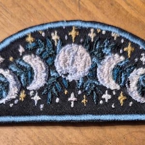 May include: Embroidered patch featuring a crescent moon design with a central full moon and surrounding stars. The patch has a black background with blue and gold accents, and a light blue border.