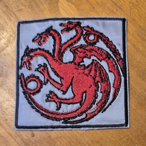 May include: A red embroidered patch with a three-headed dragon design. The dragon is surrounded by a black outline and is on a gray background.