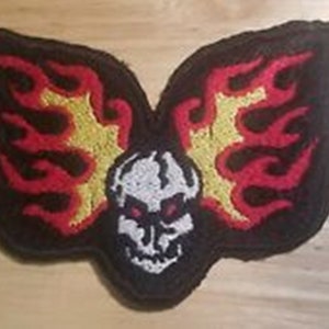 May include: A black embroidered patch with a skull in the center and flames on either side. The skull is white with black eyes and a red tongue. The flames are red and yellow.