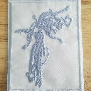 May include: A square, white embroidered patch with a light blue design of a stylized figure. The figure has outstretched arms and a tail. The patch has a light blue border.