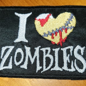 May include: A black embroidered patch with a white "I" and a stitched heart with red and yellow accents. The text "ZOMBIES" is embroidered in white below the heart.