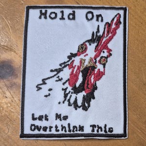 May include: Embroidered patch with the text "Hold On, Let Me Overthink This." The patch features a stylized, abstract design in red, black, and gold on a light blue background, framed by a black border.
