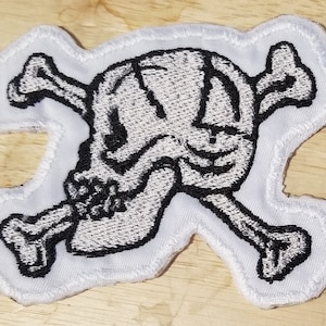 Op de afbeelding: Witte geborduurde patch met een zwart schedel- en kruisbeenderontwerp. De schedel heeft een gedetailleerd gezicht met een brede glimlach.