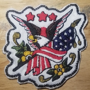 May include: A white embroidered patch with a bald eagle holding an American flag. The eagle is surrounded by three red stars and green leaves with yellow flowers.