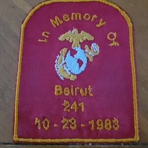 May include: A red embroidered patch with a gold border. The patch features the Marine Corps emblem, a globe, and an eagle. The text "In Memory Of" is above the emblem, and the text "Beirut 241 10-23-1983" is below the emblem.