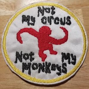 May include: A white round embroidered patch with a yellow border. The patch features a red monkey silhouette and the text "Not My Circus Not My Monkeys".