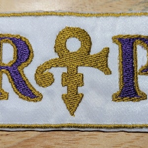 May include: A white embroidered patch with a gold border. The patch features the letters "R" and "P" in purple with gold outlines. A gold stylized symbol is between the letters.