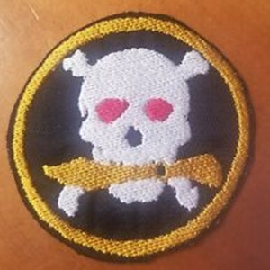 May include: Embroidered patch featuring a white skull with red eye sockets and a yellow knife through its teeth, set against a black background. The patch is circular, with a yellow border.