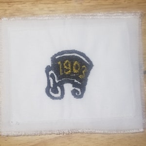 May include: White fabric patch with black embroidery of a helmet with the year 1903 in gold lettering.