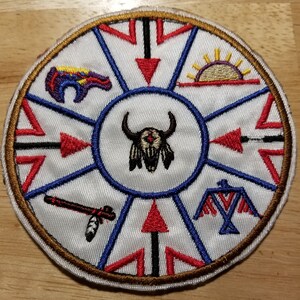 May include: A white circular patch with a brown border, featuring a Native American design. The center depicts a buffalo skull with feathers. The surrounding design includes a sun, a bird, an arrow, a pipe, and a bear.