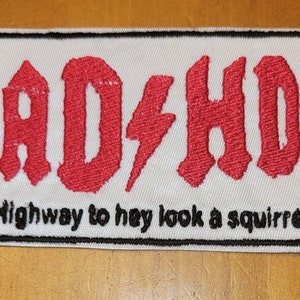 Peut inclure: Patch brodé blanc avec une bordure noire. Le patch présente le texte "AD/HD" en rouge avec un éclair entre les lettres. Sous le texte se trouve la phrase "Highway to hey look a squirrel!"