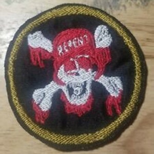 Puede incluir: Parche bordado con una calavera y huesos cruzados, un pañuelo rojo con la palabra "REPENT" y un borde dorado. La calavera es blanca con detalles rojos, sobre un fondo negro.