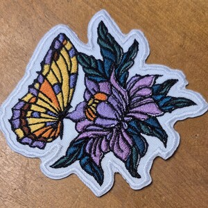 Pode incluir: Patch bordado com uma borboleta com asas amarelas, laranja e roxas, perto de uma flor roxa com folhas verdes e azuis. O patch tem uma borda branca, para roupas ou acessórios.