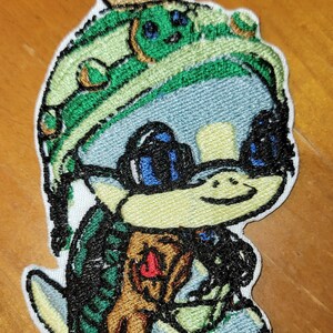 May include: Embroidered patch of a cartoon turtle wearing a green hat with a brown brim, sunglasses, and a brown shell. The turtle is smiling and holding a green leaf in its right hand.