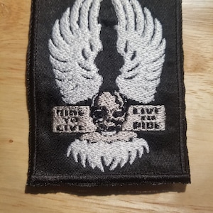May include: Black embroidered patch with a skull and wing design, and the words "Ride to Give". The patch has a black border and is designed to be sewn onto clothing or accessories. The design is in shades of grey and white.