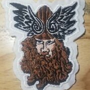 Może przedstawiać: Naszywka haftowana przedstawiająca stylizowaną postać z skrzydłami i brodą. Postać ma brązową brodę i włosy, a skrzydła są czarne z misternymi wzorami.