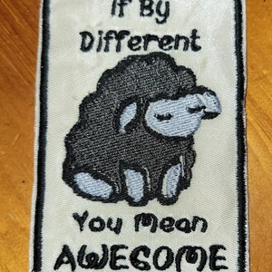 Może przedstawiać: Biała naszywka haftowana z czarną owcą i tekstem "If by different you mean awesome".