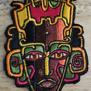 以下が含まれることがあります： 幾何学模様でスタイリッシュな人間の顔を表現したカラフルな刺繍パッチ。顔は主に茶色で、黄色、オレンジ、赤、緑、黒のアクセントが付けられています。このパッチは、衣服、バッグ、その他のアクセサリーにユニークなタッチを加えるのに最適です。