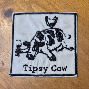 Puede incluir: Parche cuadrado con borde negro y fondo crema. Presenta una ilustración de vaca en blanco y negro con un pollo en la espalda. El texto "Tipsy Cow" está bordado en negro debajo.