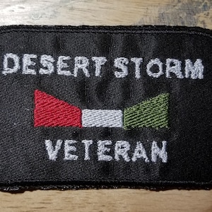 Może przedstawiać: Czarna naszywka haftowana z białym tekstem, który brzmi "DESERT STORM VETERAN". Naszywka przedstawia flagę w kolorze czerwonym, białym i zielonym.