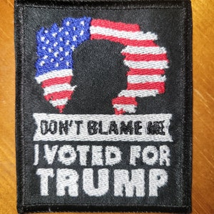 May include: Black embroidered patch with a silhouette of the United States flag and the text "Don't Blame Me I Voted For Trump".