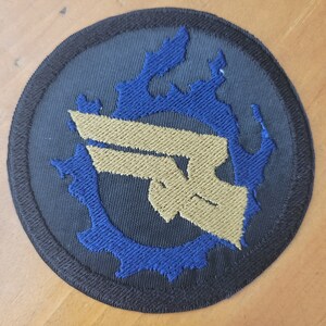 Könnte beinhalten: Runder, gestickter Aufnäher mit einem goldenen Emblem auf dunkelblauem Hintergrund, umgeben von einem blauen, flammenartigen Design. Der Aufnäher hat einen schwarzen Rand.
