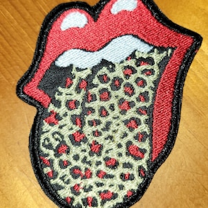 Puede incluir: Parche bordado con una lengua roja con contorno negro y un diseño de estampado de leopardo en el interior. La lengua sobresale y tiene un contorno blanco alrededor de la parte superior.