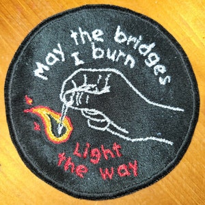 May include: Black embroidered patch with a white hand holding a match that is lighting a flame. The text reads "May I burn the bridges light the way."